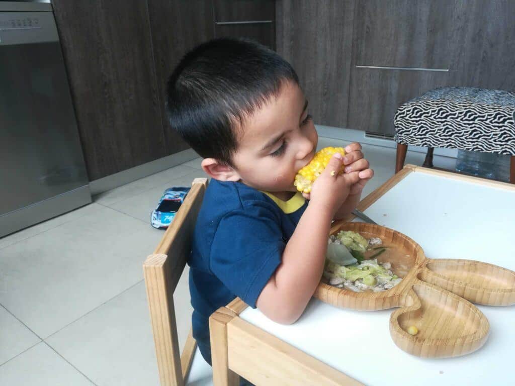 A boy seated in a table eating corn with other vegetables on his plate.